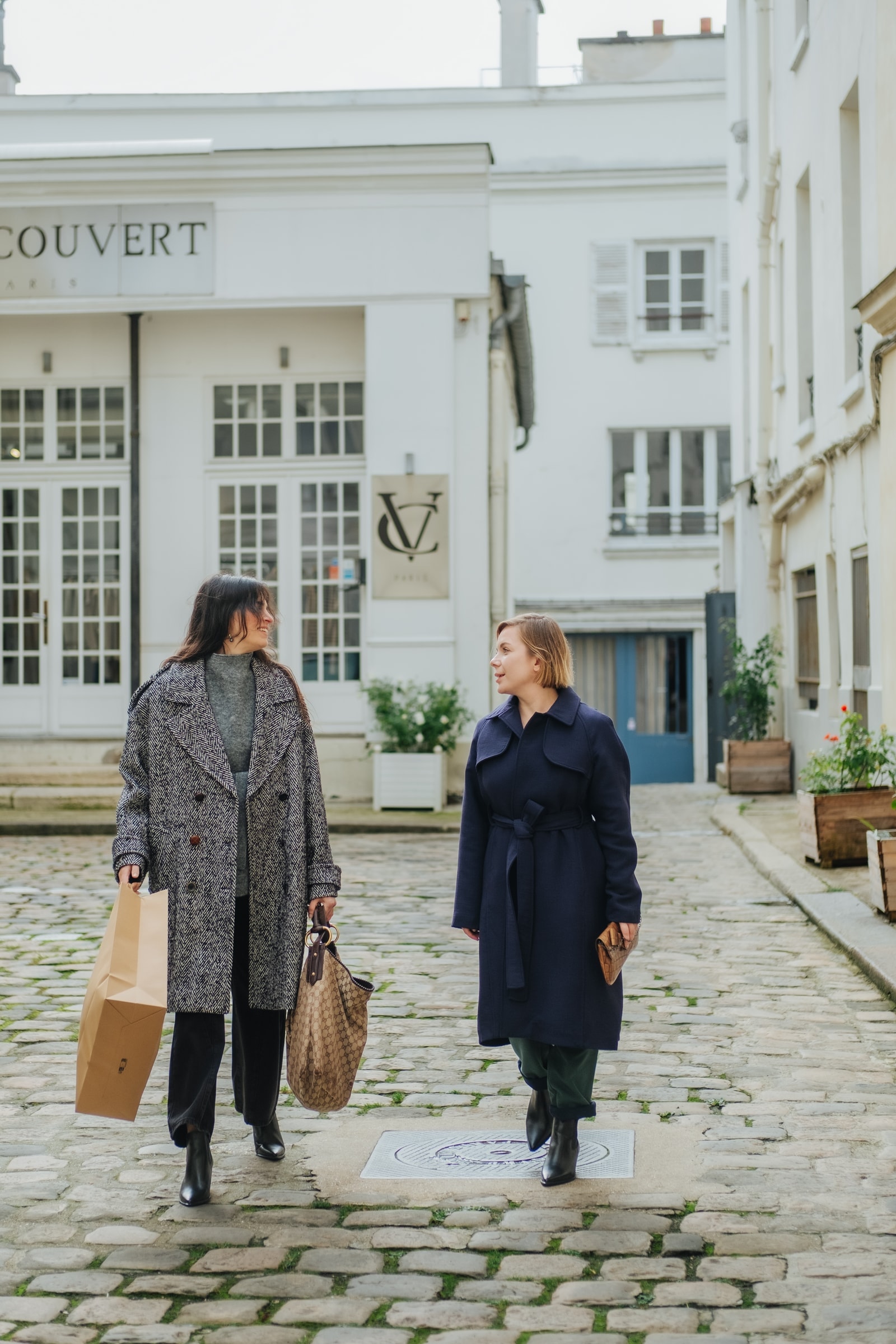 Two clients walking with luxury shopping bags in front of a Paris boutique on cobblestones